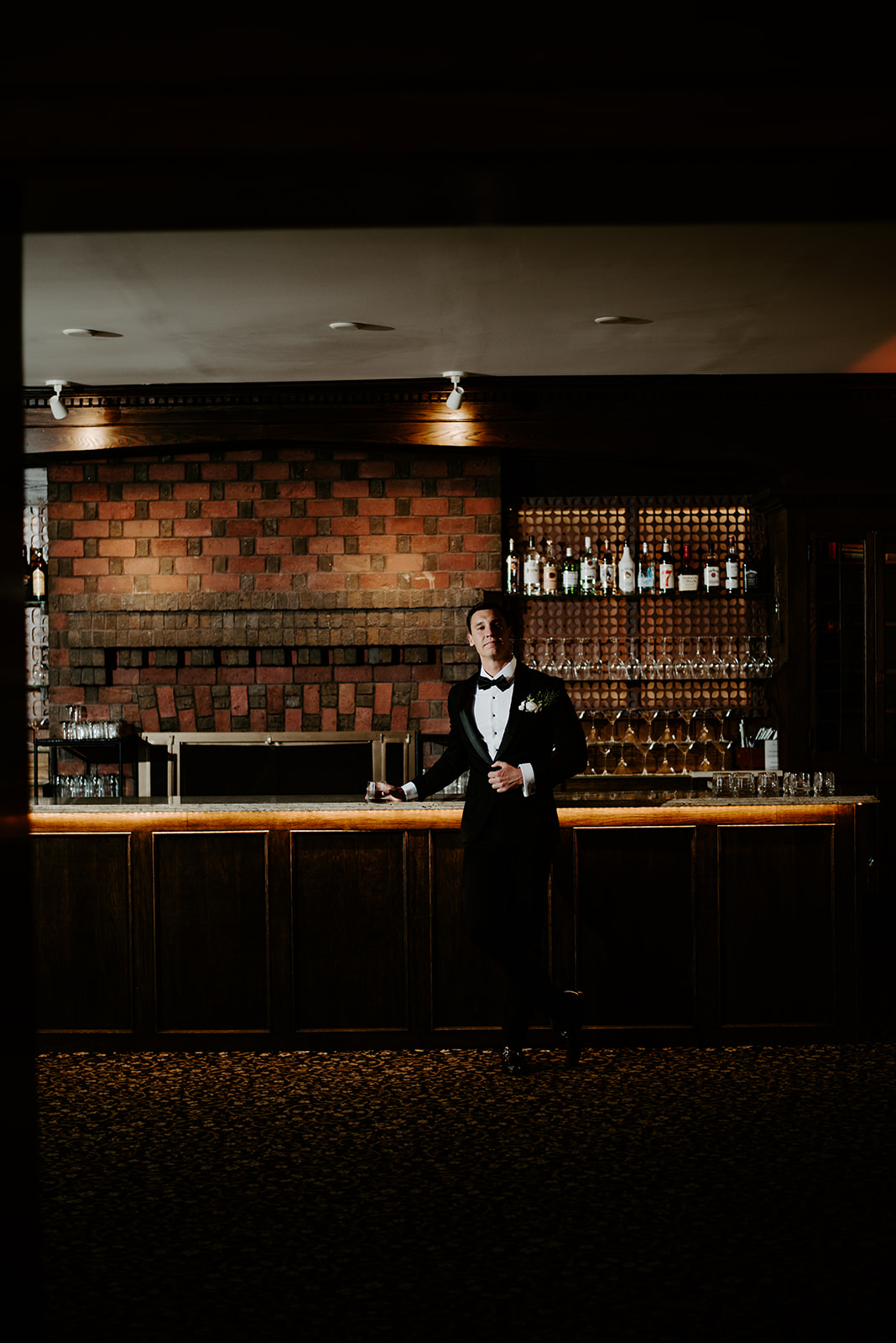 Groom in a tuxedo posing confidently with a drink at the dimly lit brick bar.